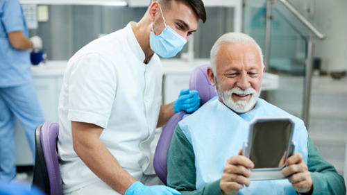 Smiling older male patient in dental chair reviewing results with masked dentist nearby